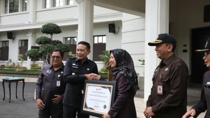 Wali Kota Malang, Dr. Ir. Wahyu Hidayat, M.M., menyerahkan piagam penghargaan WBBM tersebut kepada Kepala Dispendukcapil Kota Malang, Ir. Dahliana Lusi Ratnasari, M.M. (Sumber Prokompim).